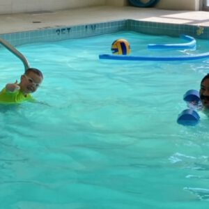 Child giving a thumbs up while swimming in a pool with a swim instructor, featuring pool toys and a bright, inviting atmosphere, reflecting the personalized swimming lessons at Aqua Life Swim Academy.