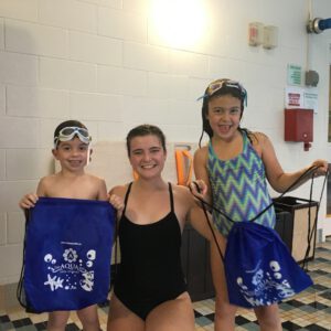 Two children holding Aqua Life Swim Academy bags with a smiling instructor in a swim facility, showcasing a fun and engaging atmosphere for swimming lessons.
