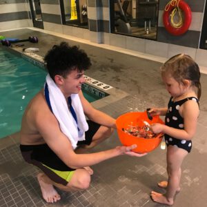 Swimming instructor engaging with a young girl in a swimsuit, sharing a bowl of swimming toys, in a pool environment at Aqua Life Swim Academy.
