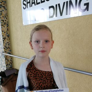 Young girl wrapped in a towel holding a swimming certificate, standing in front of a "Shallow End Diving" sign, celebrating her swimming achievement at Aqua Life Swim Academy.