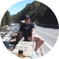 Person sitting on a stone wall by a river, wearing a black t-shirt and cap, surrounded by trees and a road, reflecting a love for the outdoors and active lifestyle.